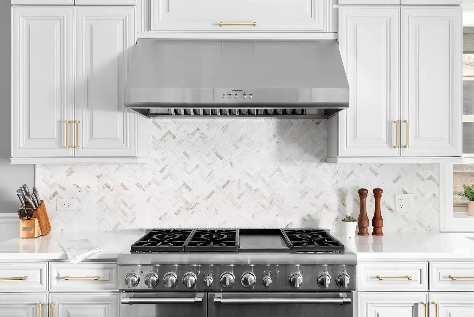 A modern gas stove in a white kitchen with a stainless steel range hood, elegant herringbone backsplash, and white marble countertops.