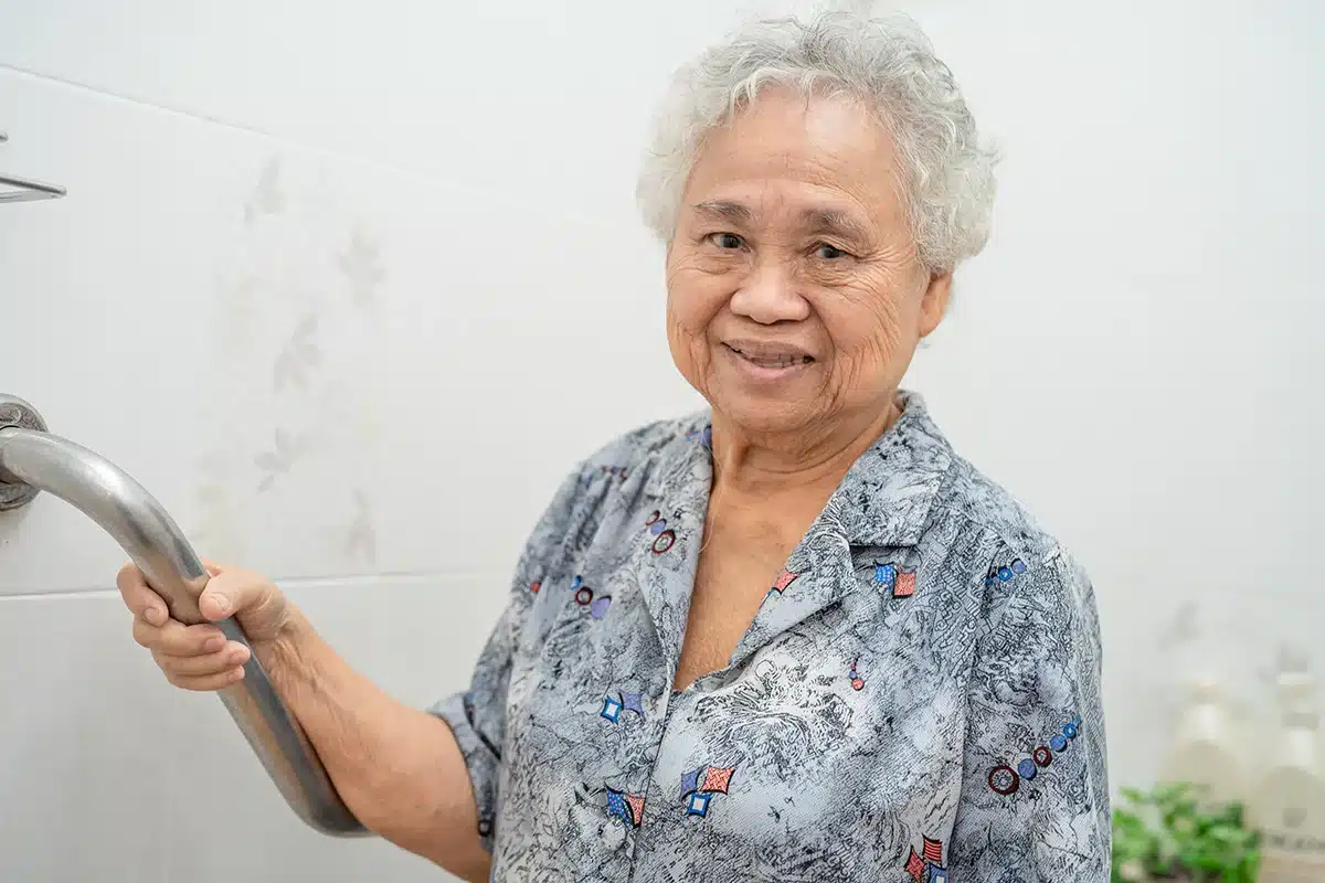 Elderly woman with a content smile holding onto grab bars in a bathroom, illustrating senior-friendly safety features.
