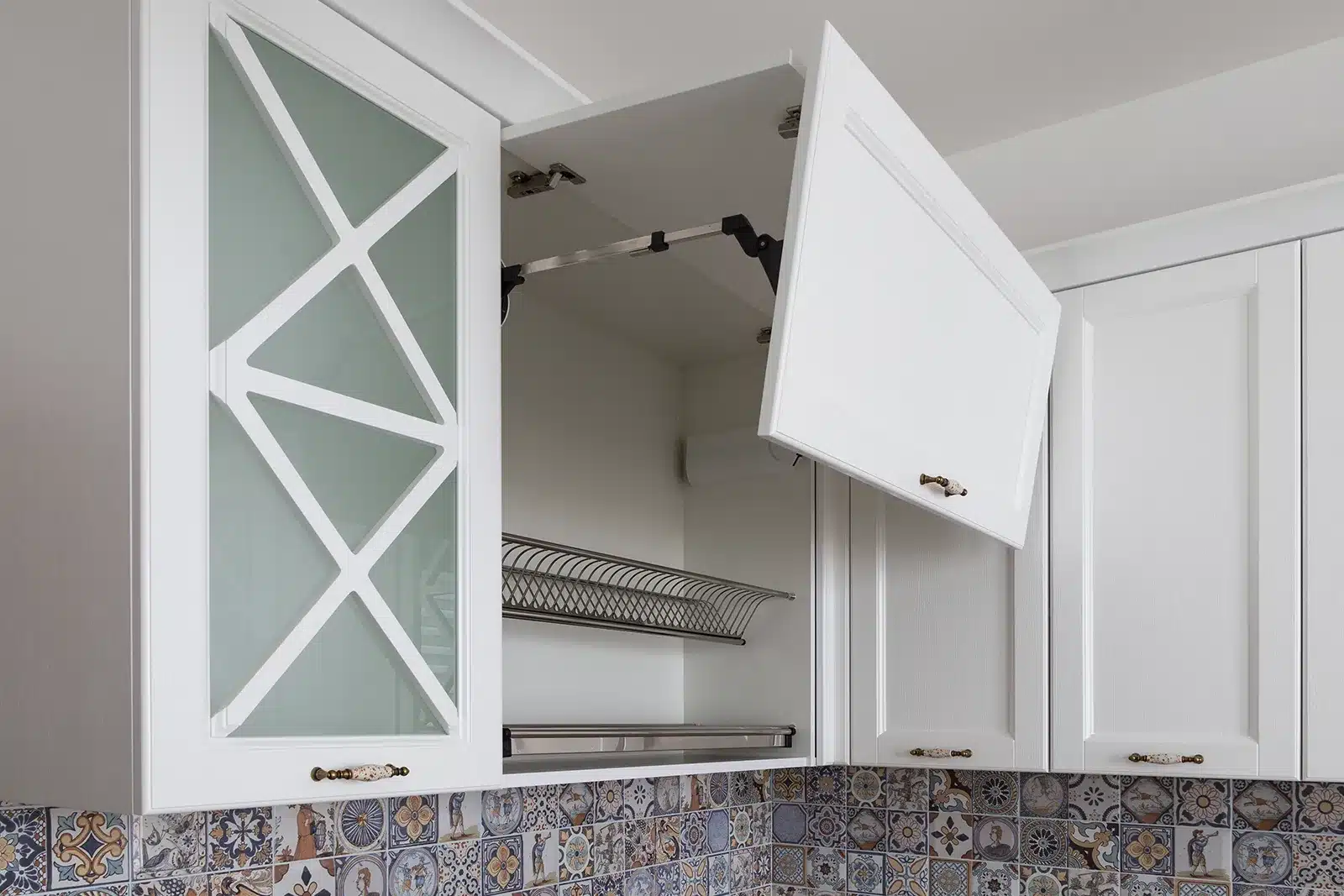 White kitchen upper cabinets with top hinges opening upwards, featuring frosted glass with a geometric pattern, over a decorative tiled backsplash.