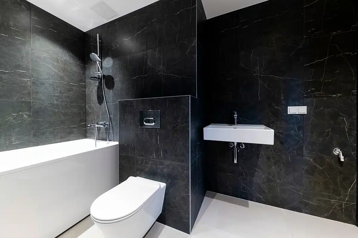 Modern bathroom with dark marble tiles, wall-mounted sink, and in-wall faucet next to a white bathtub and toilet.