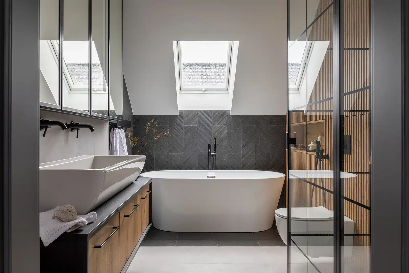 Modern bathroom remodel in Federal Way with a white freestanding bathtub, dark grey tiled walls, wooden vanity with dual sinks, and a glass-enclosed shower area.