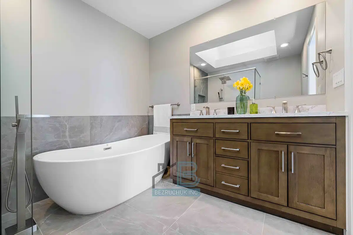 Modern Tacoma bathroom remodel featuring a white freestanding bathtub, glass shower partition, and a wooden vanity with marble countertop, accented with fresh yellow flowers and a large mirror illuminating the space.