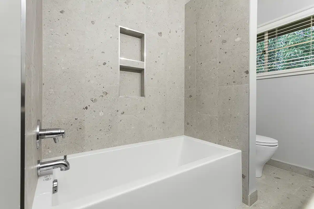 This is a contemporary bathroom on Mercer Island, showcasing a pristine white bathtub. Complemented by terrazzo wall tiles and niche shelving, it also offers a lush-green view through the window.